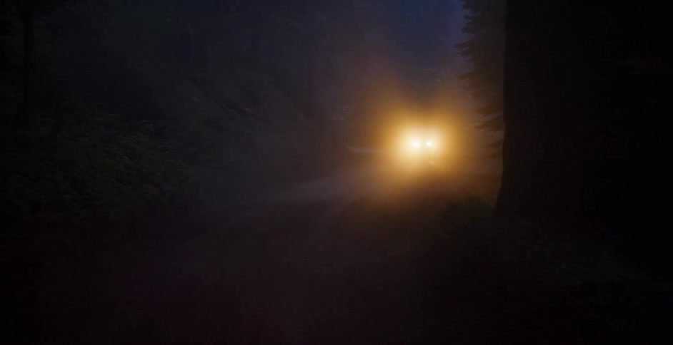 Pan shot of a car's headlights cutting through dense fog as it drives down a spooky forest road at night