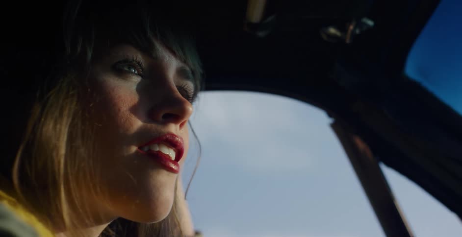 A cinematic close-up shows a thoughtful woman with red lipstick gazing out of a car window during a golden hour drive