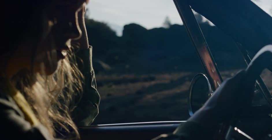 A silhouetted woman in a classic car passenger seat enjoys the scenery on a country road during a golden hour drive in slow motion
