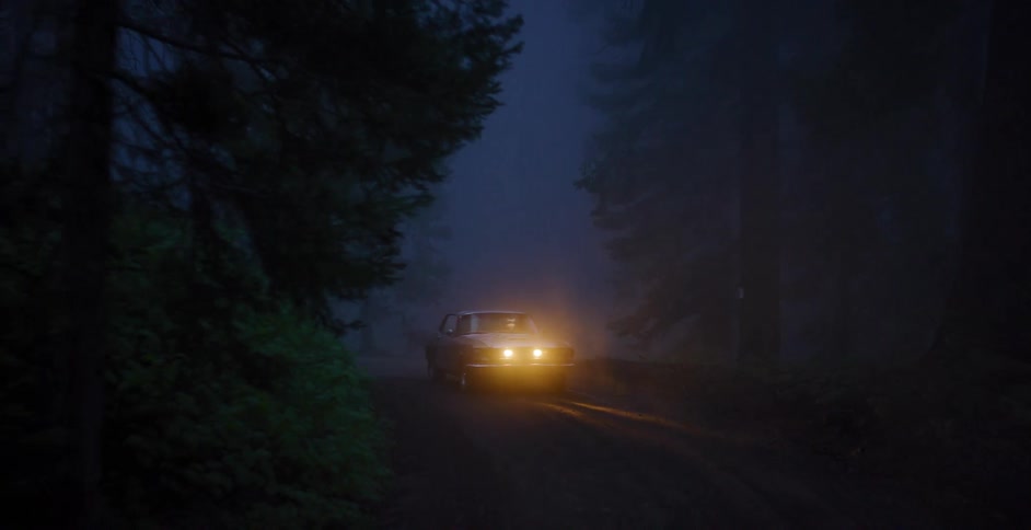 A vintage car with glowing headlights is parked on a dirt road in a mysterious, foggy forest at night