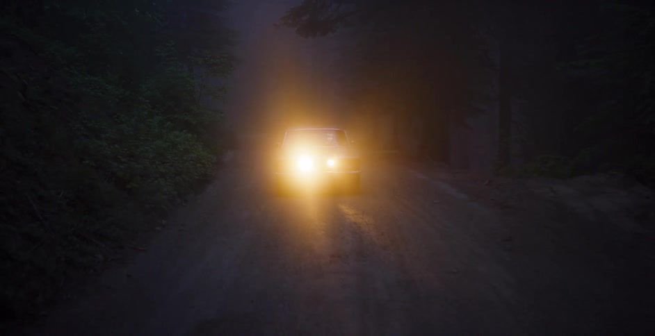 A classic American muscle car emerges from the thick fog, its headlights illuminating a dark and mysterious forest road at night