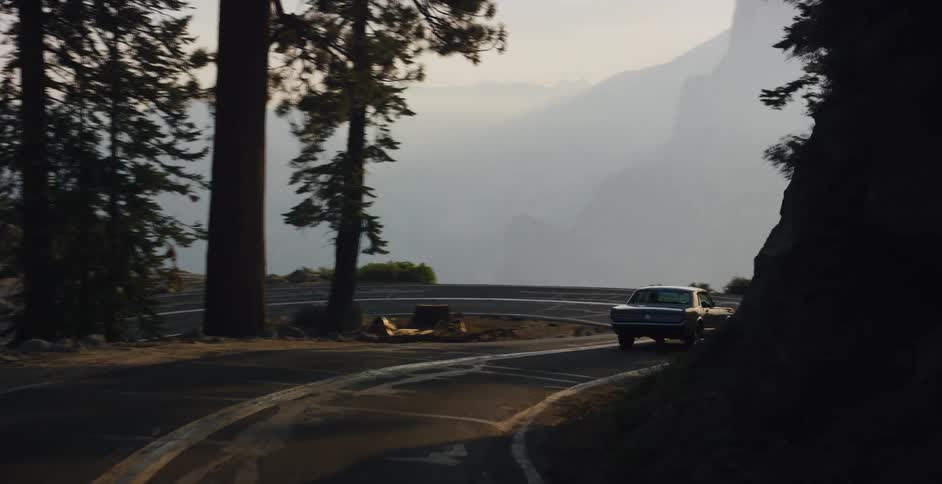 A classic car navigates a winding road through a sunlit forest, opening up to a spectacular hazy vista of Yosemite National Park