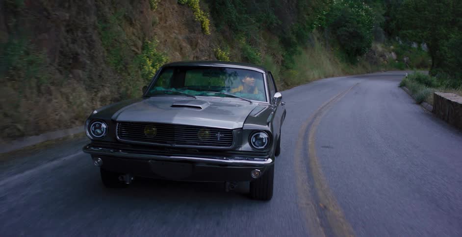 Front-facing shot of a woman confidently driving a classic grey muscle car down a winding country road