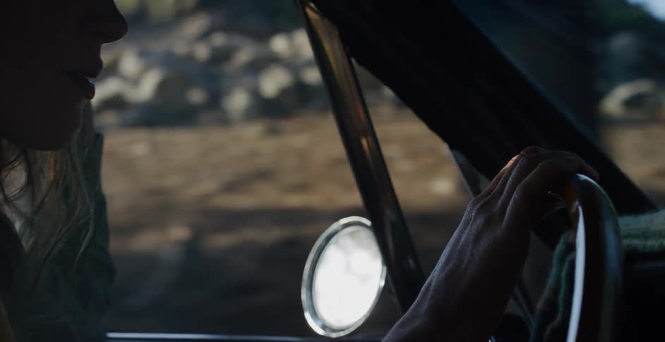 A woman's hands steer a vintage car down a country road as sunlight flickers through the window