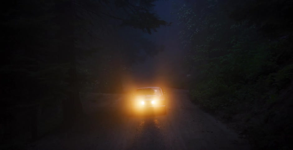 A car's bright headlights pierce the dense fog on a dark, mysterious forest road at night as the camera slowly pulls back
