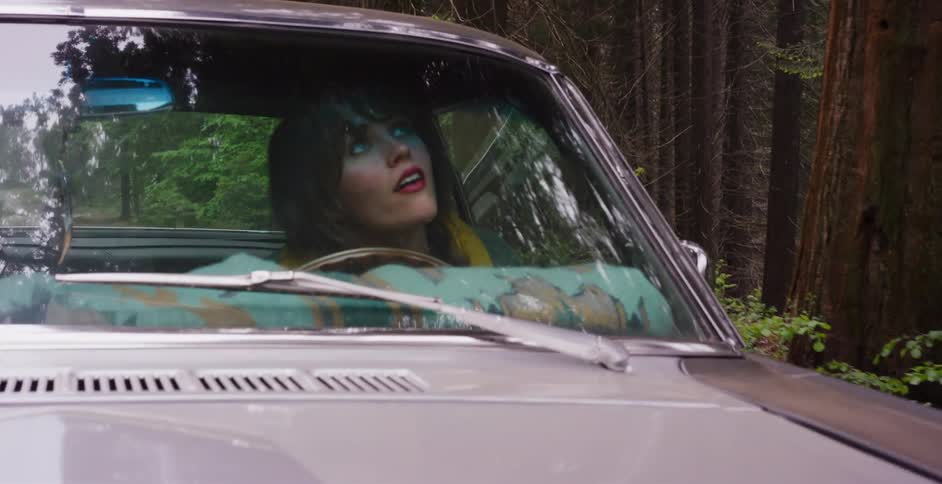 A young woman in a classic car gazes up in wonder at the towering redwood trees in a lush, dense forest