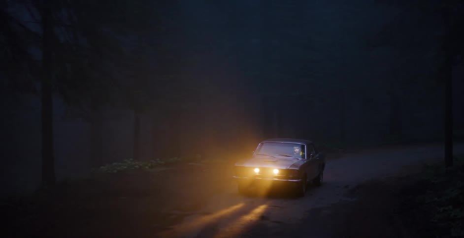 A classic American muscle car with bright headlights on drives along a dark, wet, and foggy road through a spooky forest at night