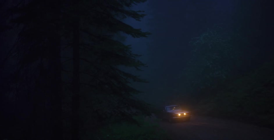 A lone car with its headlights on illuminates a dark, foggy road deep within a forest at night