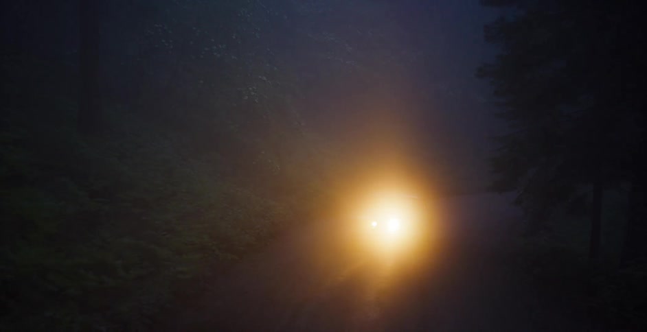 A classic car with bright headlights on sits parked on a remote, foggy forest road at night
