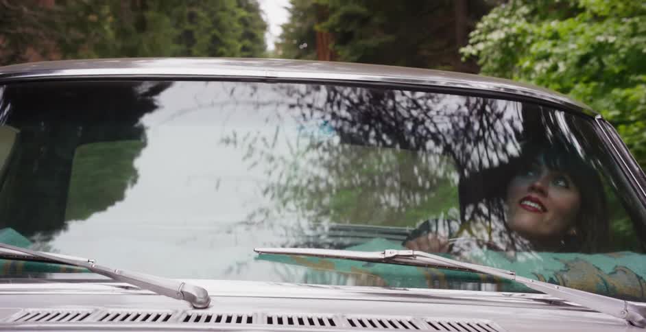 A happy woman in the passenger seat of a classic car looks out at the forest scenery reflecting on the windshield