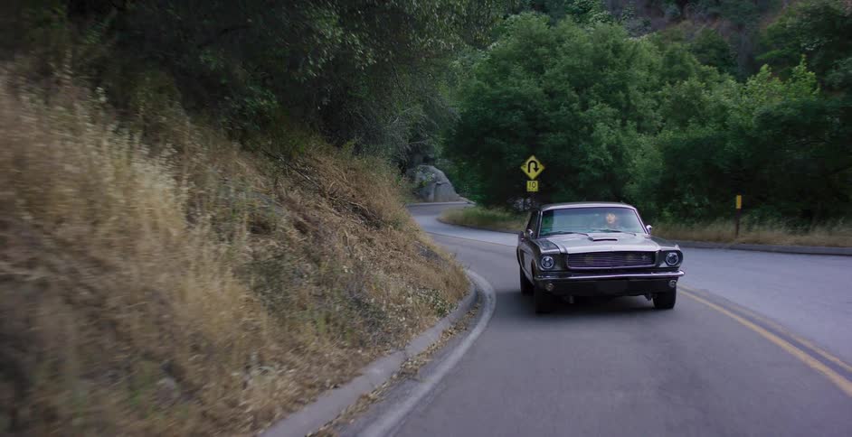 A classic silver Mustang navigates the curves of a scenic, tree-lined mountain road