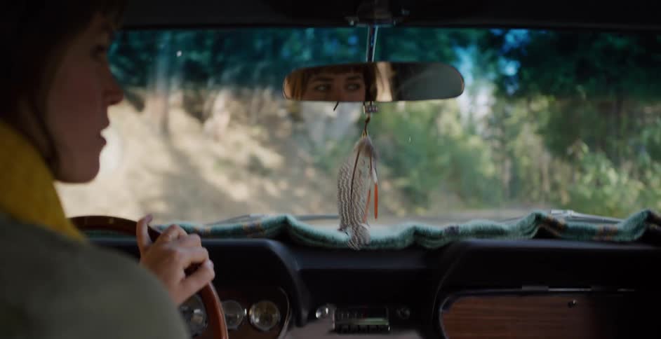 A woman takes a scenic drive through a sunny forest in her classic car, with a feather swaying from the rearview mirror