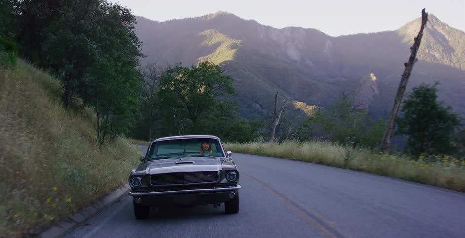 A woman takes a scenic drive through the mountains in a classic dark Ford Mustang at sunset