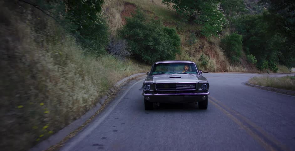 A woman drives a classic grey Mustang in slow motion down a winding, tree-lined road in the mountains