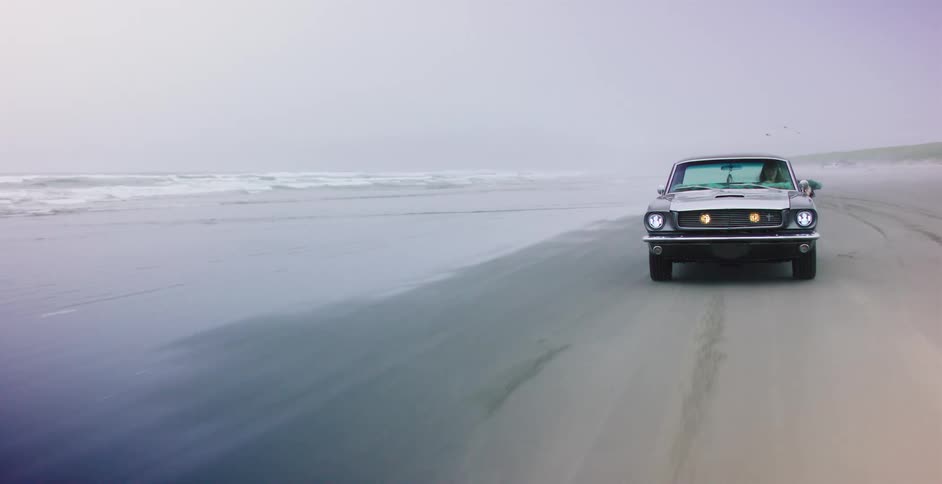 A classic muscle car cruises down a foggy beach with headlights on, driving beside the ocean waves