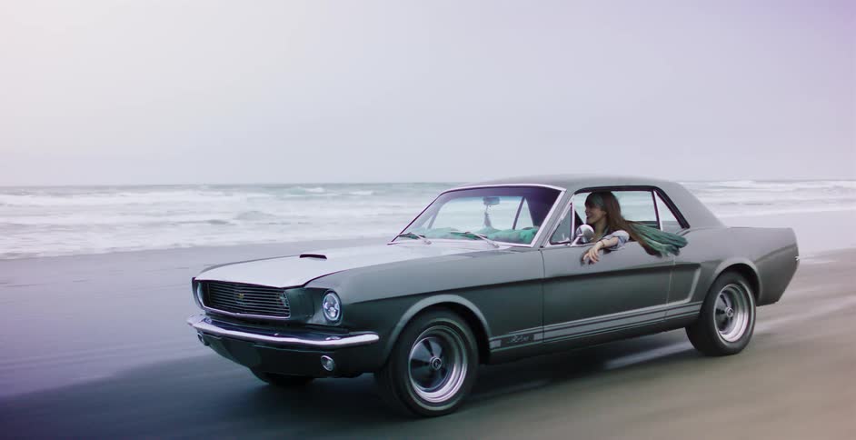 A woman takes a scenic drive in a classic grey Ford Mustang along the sandy shoreline of an overcast beach