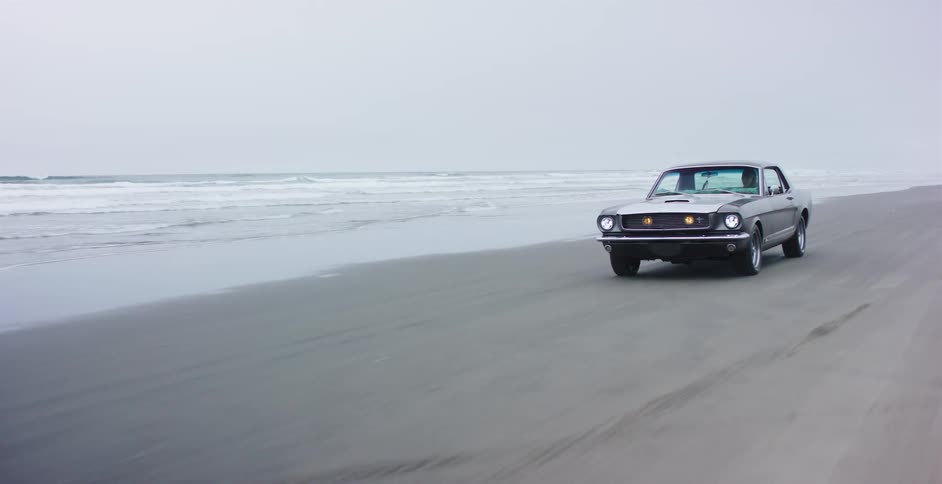A classic silver Ford Mustang cruises along the wet sand of an ocean beach on a foggy, overcast day