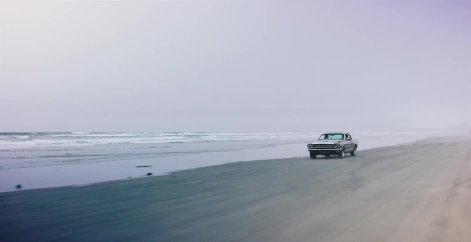 A classic muscle car cruises down a vast, foggy beach as waves from the ocean roll onto the sandy shore