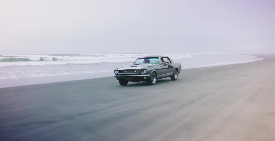 A vintage grey muscle car cruises along a foggy beach with the ocean waves in the background