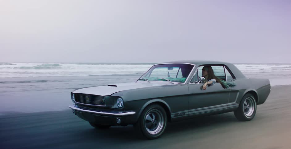A stylish woman enjoys a solo drive in a classic grey muscle car along the coastline on a cool, overcast day