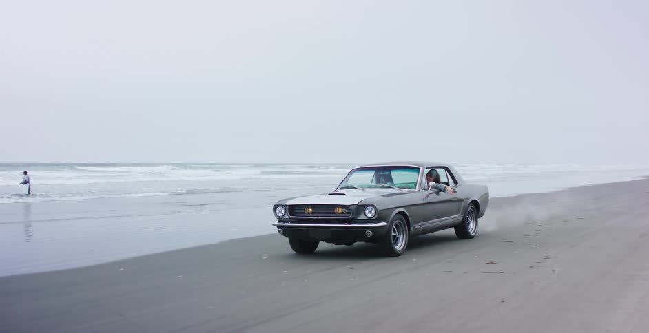 A classic silver Ford Mustang kicks up sand in slow motion as it cruises along the ocean's edge on a foggy day