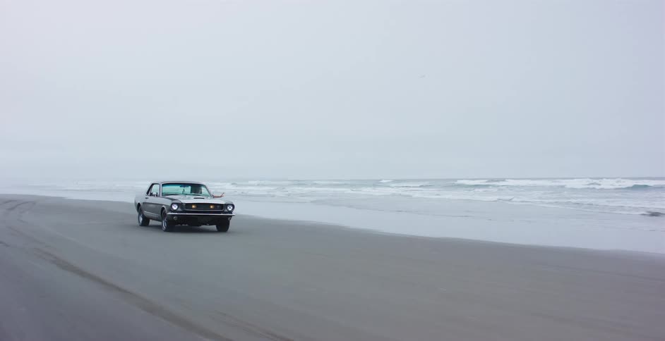 A vintage Ford Mustang drives along a vast, misty beach next to the crashing waves of the ocean
