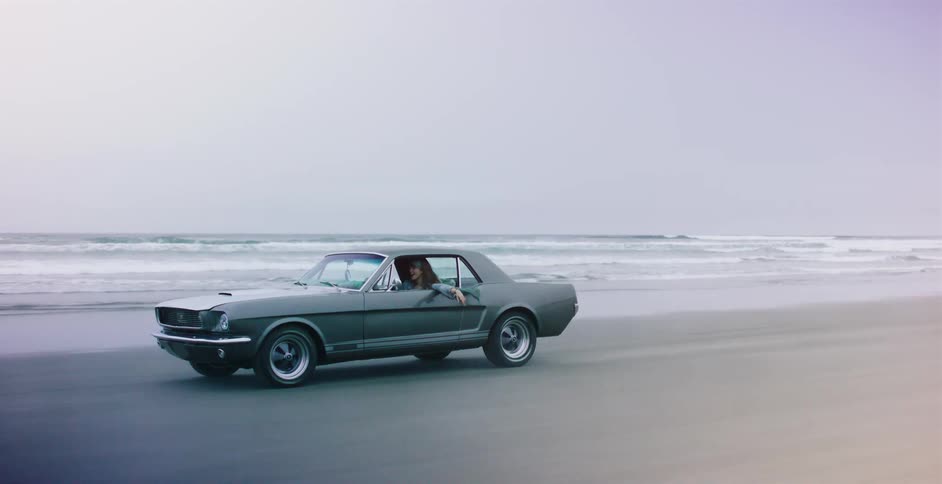 A couple takes a scenic drive in a classic gray Ford Mustang along a wide, foggy beach next to the ocean