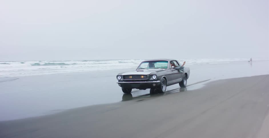 Tracking shot of a cool vintage Mustang cruising down the beach next to the ocean waves on a foggy day