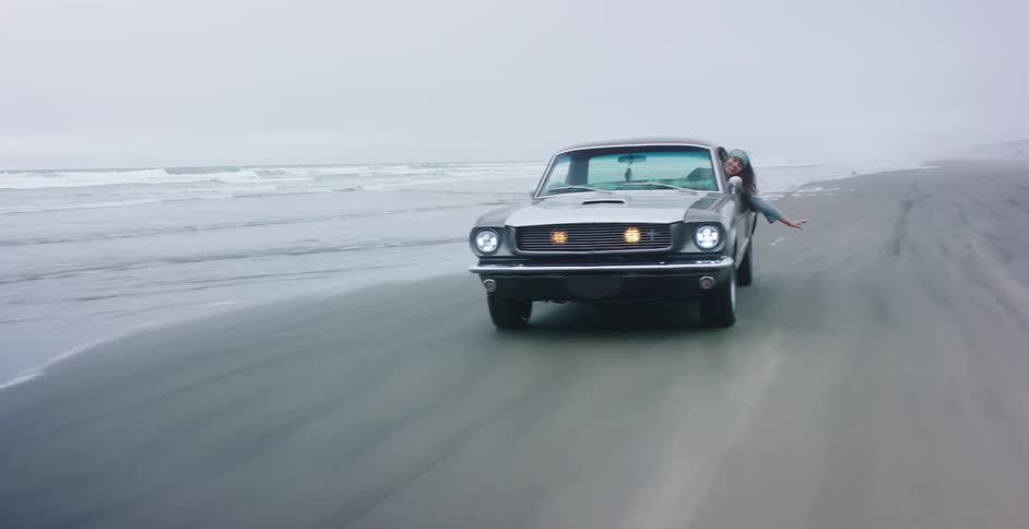 A carefree woman leans out of a classic Ford Mustang as it drives along the ocean shoreline on a foggy day