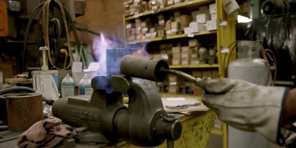 A man with a torch directs the flames on a piece of metal held in a vise