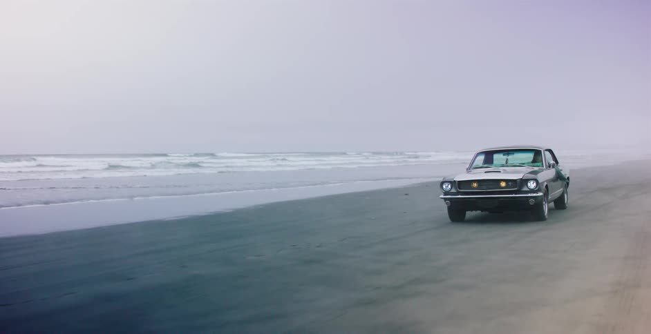 A stylish classic muscle car drives along the ocean shoreline on a foggy, cool day