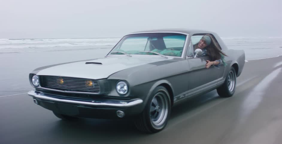 A happy young woman with long hair drives a classic silver Mustang along the ocean shoreline on an overcast day