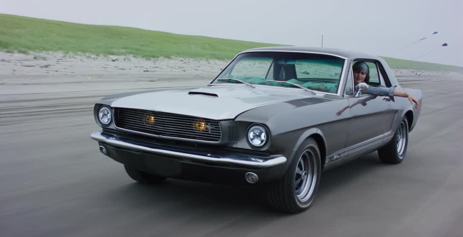 Carefree woman with her arm out the window drives a classic silver Mustang along the beach on an overcast day
