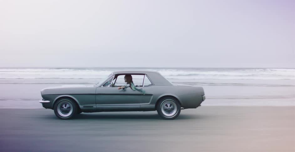 Side profile of a woman driving a classic grey Mustang down the beach next to the ocean on a foggy day