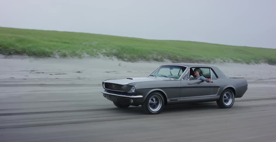 A woman enjoys a scenic drive in her classic Ford Mustang along a sandy beach on an overcast day