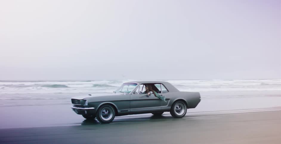 A woman drives a classic grey Ford Mustang along the wet sand of the beach next to the ocean waves on a cloudy day