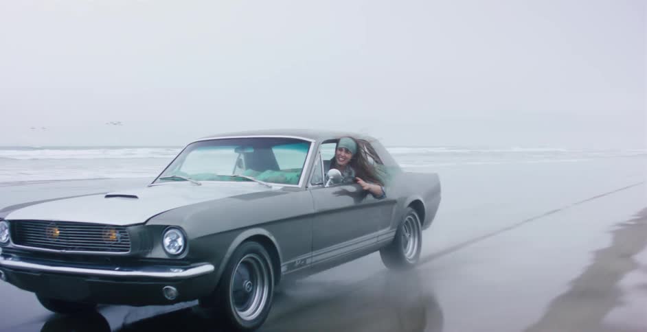 A joyful woman leans out the window of a classic Ford Mustang as it drives along a wet, sandy beach in slow motion