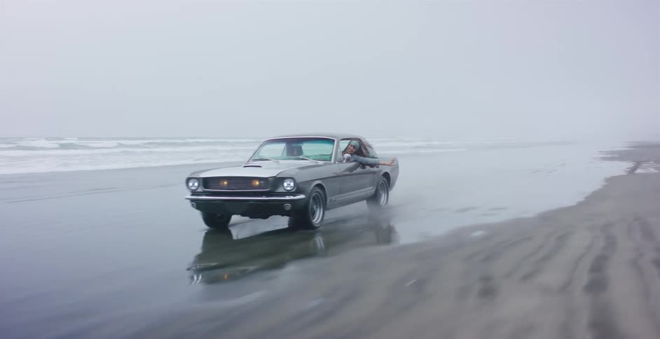 A classic grey Ford Mustang drives in slow motion along a foggy beach as a woman leans out the window, enjoying the ride