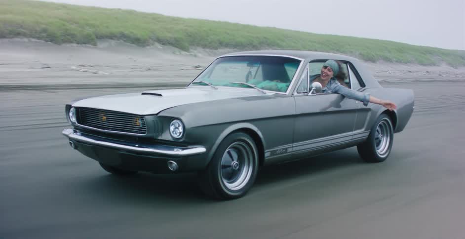 A happy young woman leans out of a classic Ford Mustang as it drives along a sandy beach on a cool, overcast day