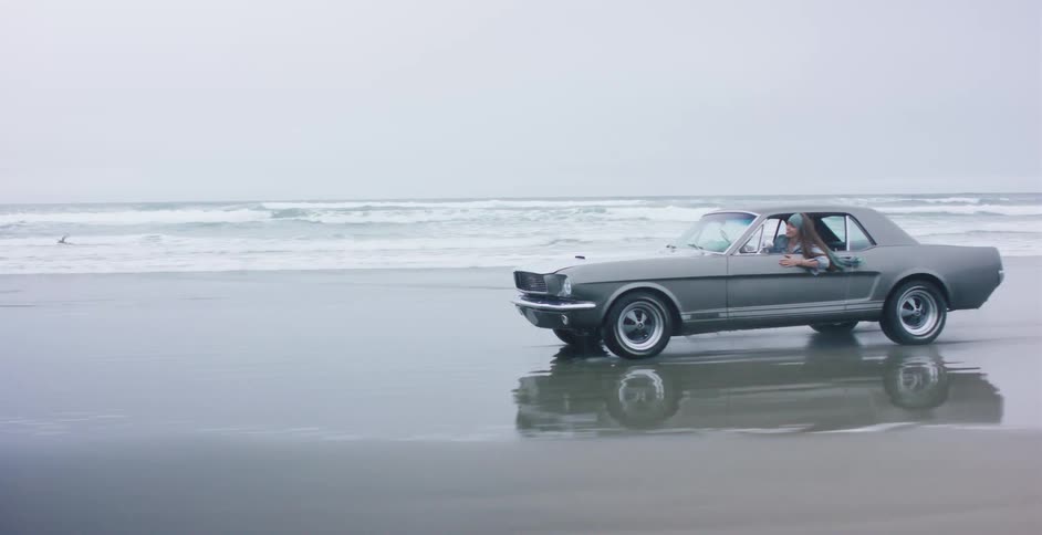 A young woman enjoys a ride in a classic Ford Mustang as it drives along the shoreline on an overcast day