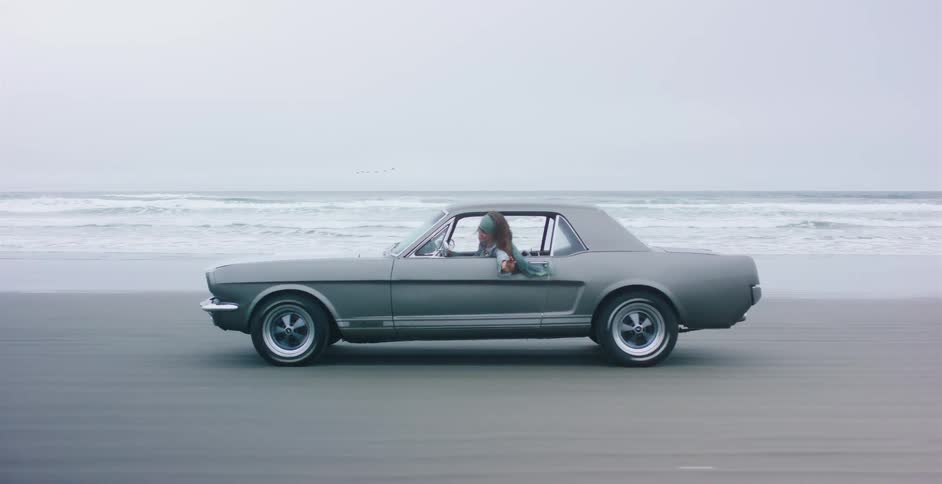 A stylish woman drives a classic grey Ford Mustang along the shoreline on an overcast day