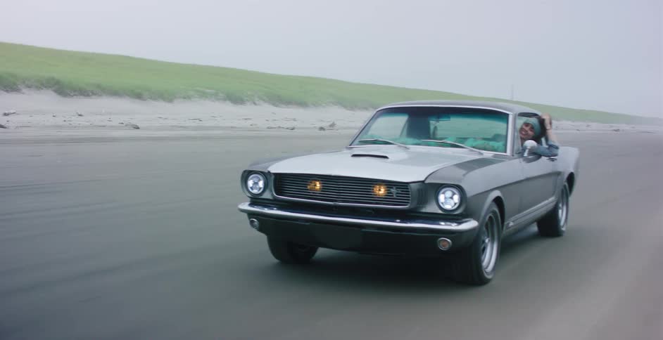 A carefree woman smiles while leaning out the window of a classic Ford Mustang cruising down a foggy beach