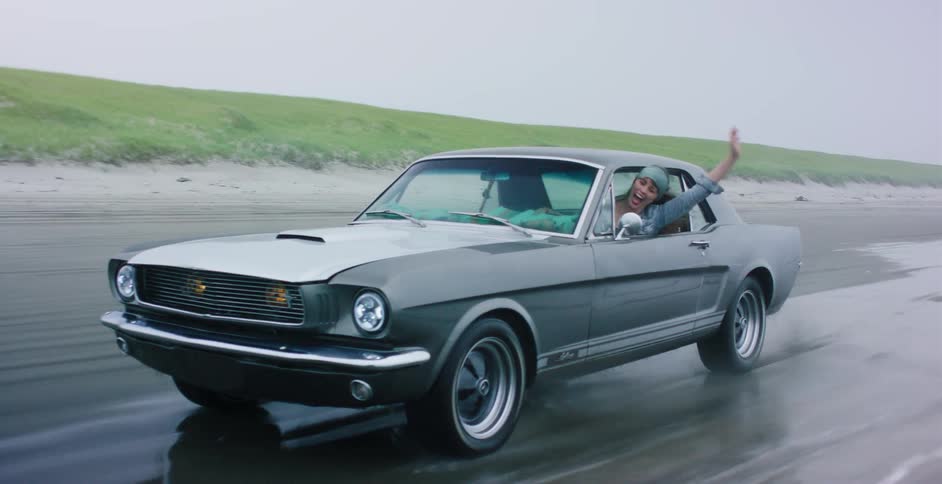 A carefree woman joyfully drives a classic silver Mustang along a wet sandy beach on a foggy day, with her arm out the window in slow motion