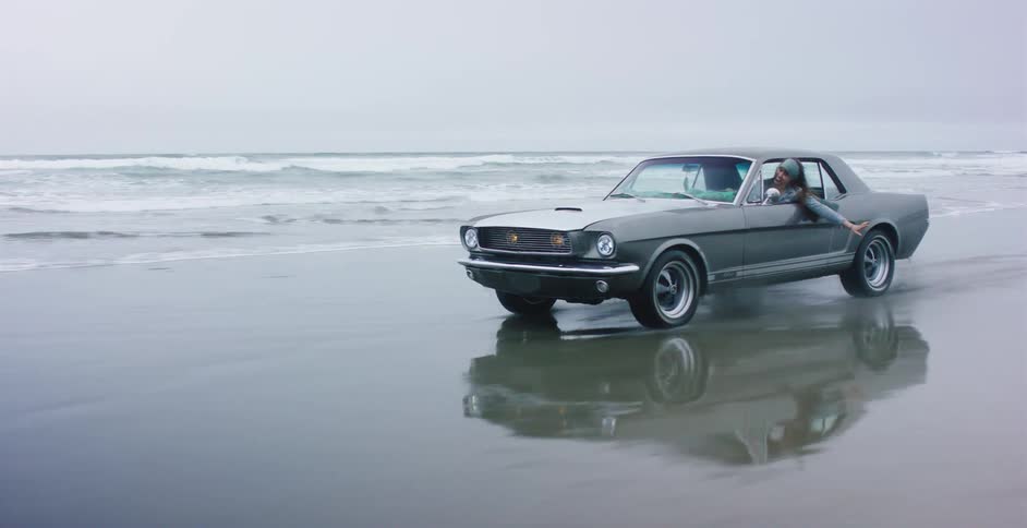 A carefree woman hangs out the window of a classic Ford Mustang as it drives along the reflective, wet sands of an ocean beach in slow motion