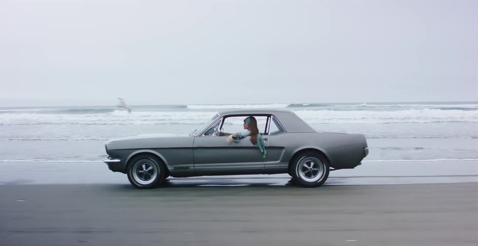 A woman in a classic grey Mustang enjoys a slow motion drive along an overcast beach as seagulls fly by