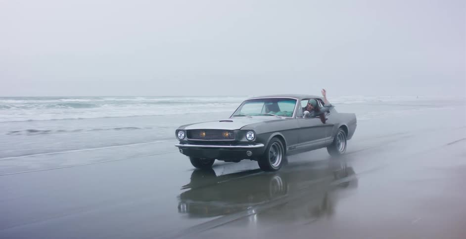 A joyful woman with long, flowing hair leans out the window of a classic grey Mustang as it drives along a wet, reflective beach in slow motion