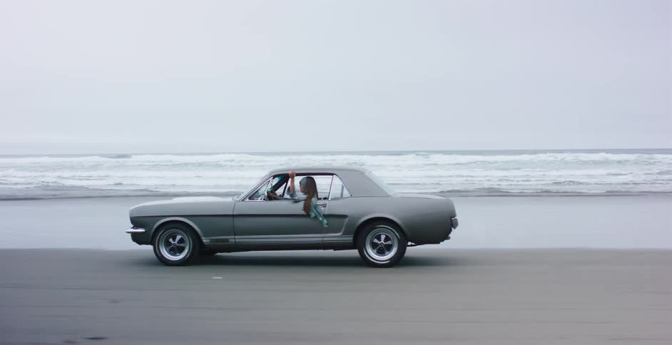 A woman drives a classic grey muscle car down the wet sand on an ocean beach on an overcast day