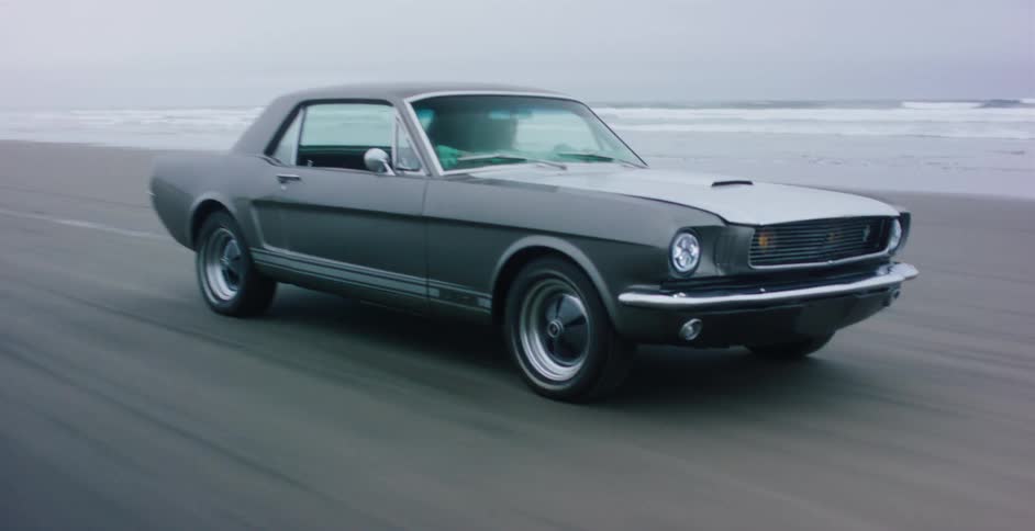 A classic gray Ford Mustang cruises down an expansive, sandy beach next to the ocean on an overcast day
