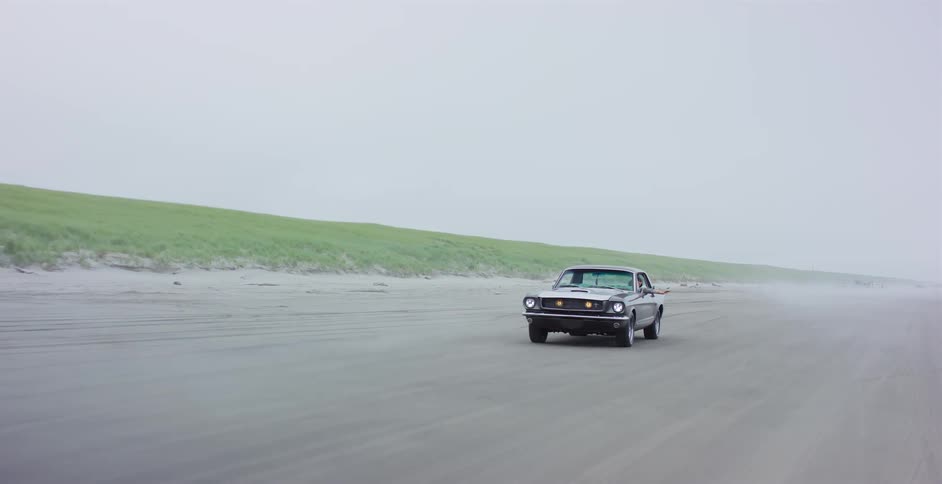 A woman enjoys the freedom of driving a classic Ford Mustang along a wide sandy beach on a foggy day