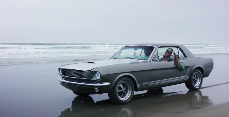 A free-spirited woman with long hair drives a classic grey Ford Mustang along the shoreline of an overcast beach